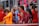 A group of young female athletes in sports attire are gathered together, some wearing red jackets or maroon jerseys with the logo of a soccer academy. They appear to be standing in front of a wire fence with spectators in the background, dressed in casual clothing.