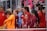 A group of young female athletes in sports attire are gathered together, some wearing red jackets or maroon jerseys with the logo of a soccer academy. They appear to be standing in front of a wire fence with spectators in the background, dressed in casual clothing.