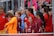 A group of young female athletes in sports attire are gathered together, some wearing red jackets or maroon jerseys with the logo of a soccer academy. They appear to be standing in front of a wire fence with spectators in the background, dressed in casual clothing.