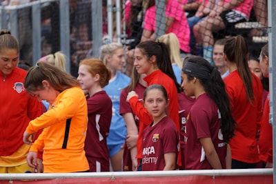 A group of young female athletes in sports attire are gathered together, some wearing red jackets or maroon jerseys with the logo of a soccer academy. They appear to be standing in front of a wire fence with spectators in the background, dressed in casual clothing.