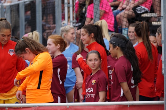 A group of young female athletes in sports attire are gathered together, some wearing red jackets or maroon jerseys with the logo of a soccer academy. They appear to be standing in front of a wire fence with spectators in the background, dressed in casual clothing.