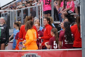 A group of female soccer players are gathered near a fence. Some are wearing red jackets, while one is in an orange jacket, and others wear maroon jerseys with numbers on the back. They appear to be interacting and conversing with one another. In the background, fans in colorful shirts are seated in bleachers, watching the scene.