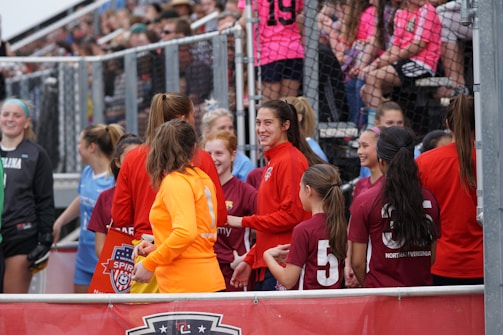 A group of female soccer players are gathered near a fence. Some are wearing red jackets, while one is in an orange jacket, and others wear maroon jerseys with numbers on the back. They appear to be interacting and conversing with one another. In the background, fans in colorful shirts are seated in bleachers, watching the scene.