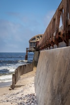 A long, rusted metal structure stretches out over a sandy shoreline toward a body of water under a clear blue sky. The structure extends from a concrete seawall lined with vertical support beams. Waves gently hit the shore, and scattered seashells and debris are visible on the sand.