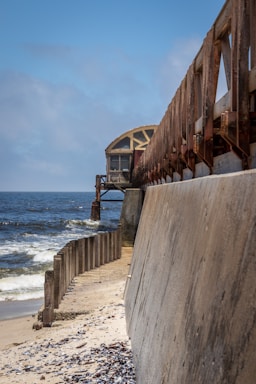A long, rusted metal structure stretches out over a sandy shoreline toward a body of water under a clear blue sky. The structure extends from a concrete seawall lined with vertical support beams. Waves gently hit the shore, and scattered seashells and debris are visible on the sand.