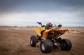 A yellow quad bike is parked on a sandy beach with tire tracks visible on the ground. The bike has a helmet placed on its seat, and there are rows of white barriers or tires in the background. The sky is overcast, creating a moody atmosphere.