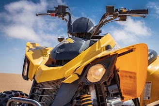 A yellow all-terrain vehicle (ATV) with rugged tires is parked in a sandy desert environment. The focus is on the front view of the vehicle, showcasing its handlebars and headlight. The background features a clear blue sky with a few scattered clouds.