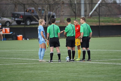A group of people stand in a circle on a soccer field. Two players, one in red and yellow and the other in light blue, are surrounded by three referees wearing green jerseys and black shorts. A soccer ball is placed on the ground in the center of the group.