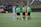 A group of people stand in a circle on a soccer field. Two players, one in red and yellow and the other in light blue, are surrounded by three referees wearing green jerseys and black shorts. A soccer ball is placed on the ground in the center of the group.