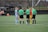 A group of people stand in a circle on a soccer field. Two players, one in red and yellow and the other in light blue, are surrounded by three referees wearing green jerseys and black shorts. A soccer ball is placed on the ground in the center of the group.