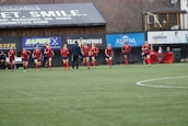 A group of soccer players in red and black uniforms are running on a green field towards the camera. A coach or trainer stands in front of them, possibly giving instructions. Advertising boards and a banner are visible in the background, along with some spectators seated at the sidelines.