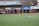 A group of soccer players in red and black uniforms are running on a green field towards the camera. A coach or trainer stands in front of them, possibly giving instructions. Advertising boards and a banner are visible in the background, along with some spectators seated at the sidelines.