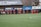 A group of soccer players in red and black uniforms are running on a green field towards the camera. A coach or trainer stands in front of them, possibly giving instructions. Advertising boards and a banner are visible in the background, along with some spectators seated at the sidelines.