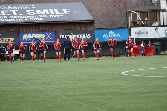 A group of soccer players in red and black uniforms are running on a green field towards the camera. A coach or trainer stands in front of them, possibly giving instructions. Advertising boards and a banner are visible in the background, along with some spectators seated at the sidelines.