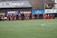 A group of soccer players in red and black uniforms are running on a green field towards the camera. A coach or trainer stands in front of them, possibly giving instructions. Advertising boards and a banner are visible in the background, along with some spectators seated at the sidelines.