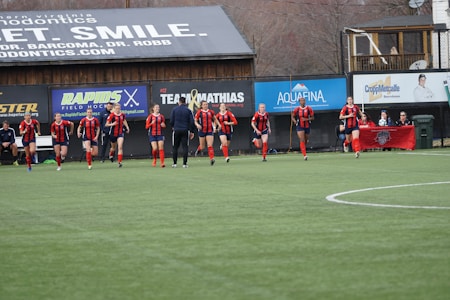 A group of soccer players in red and black uniforms are running on a green field towards the camera. A coach or trainer stands in front of them, possibly giving instructions. Advertising boards and a banner are visible in the background, along with some spectators seated at the sidelines.
