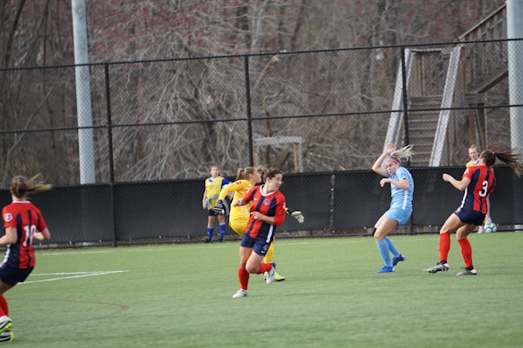 A group of female soccer players competes on a grass field. Players in red and blue uniforms are actively engaged, with one player in light blue attempting to intercept or pass the ball. The scene is outdoors, bordered by a black fencing and leafless trees in the background, suggesting a chilly or off-season time.
