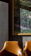 The image features two brown leather chairs placed in front of a wall with textured gray bricks. On the wall is a large framed picture depicting a lush, green forest scene with a dirt path and some indirect sunlight creating shadows. The atmosphere blends elements of interior decor with an artistic portrayal of nature.