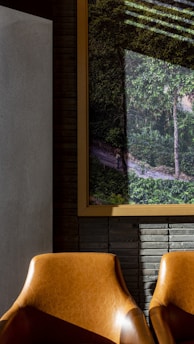 The image features two brown leather chairs placed in front of a wall with textured gray bricks. On the wall is a large framed picture depicting a lush, green forest scene with a dirt path and some indirect sunlight creating shadows. The atmosphere blends elements of interior decor with an artistic portrayal of nature.