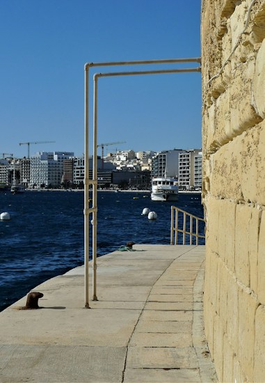 A harbor scene with a concrete walkway beside a stone wall, extending towards the water. A boat is seen on the water, with several white buoys floating nearby. In the background, there are modern buildings and construction cranes under a clear blue sky.