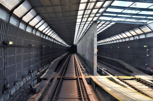 An empty railway tunnel with metal walls and a ceiling made of glass panels, allowing sunlight to stream in. The tracks run parallel, converging in the distance to a single point. The lighting fixtures are evenly spaced along the walls, and various cables and mechanical equipment line the sides.