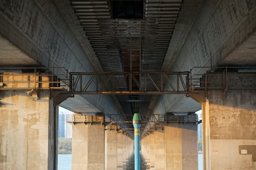 A concrete bridge viewed from underneath, showcasing large support columns and a suspended maintenance walkway running along the length of the structure. The textures of the concrete and metal elements are prominent, and there is a river visible below. The image captures the architectural details and symmetry of the bridge construction.
