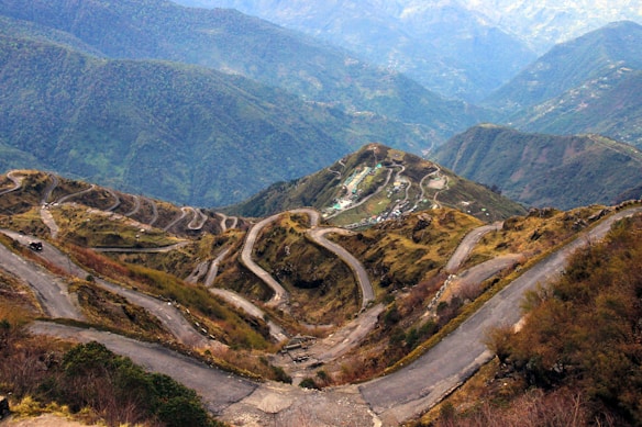 A winding mountain road cuts through lush green hills, creating a mesmerizing pattern as it descends into the valley. The landscape features rugged terrain with patches of greenery and dry vegetation, extending to distant mountain ranges under a clear sky.
