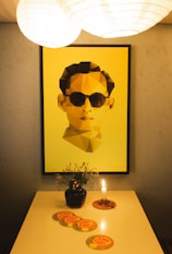 A modern art piece featuring a geometric, polygonal portrait in yellow and shades of brown hangs on a wall. Below the artwork, there is a small table with a black vase holding minimalistic floral arrangements. A single, lit candle on a circular wooden base is next to a series of colorful coasters with the text 'CHA CHA'. Above, two large white spherical lamps provide warm lighting, creating a cozy ambiance.