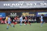 Players engage in a competitive soccer game on a field while others are seated on the bench watching attentively. The action is taking place in front of a wooden structure displaying multiple advertisements, including orthodontics, lacrosse, and field hockey.