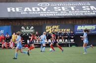 Players engage in a competitive soccer game on a field while others are seated on the bench watching attentively. The action is taking place in front of a wooden structure displaying multiple advertisements, including orthodontics, lacrosse, and field hockey.