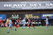 Players engage in a competitive soccer game on a field while others are seated on the bench watching attentively. The action is taking place in front of a wooden structure displaying multiple advertisements, including orthodontics, lacrosse, and field hockey.