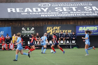 Players engage in a competitive soccer game on a field while others are seated on the bench watching attentively. The action is taking place in front of a wooden structure displaying multiple advertisements, including orthodontics, lacrosse, and field hockey.