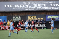 Players engage in a competitive soccer game on a field while others are seated on the bench watching attentively. The action is taking place in front of a wooden structure displaying multiple advertisements, including orthodontics, lacrosse, and field hockey.