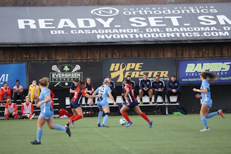 Players engage in a competitive soccer game on a field while others are seated on the bench watching attentively. The action is taking place in front of a wooden structure displaying multiple advertisements, including orthodontics, lacrosse, and field hockey.
