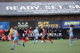 Players engage in a competitive soccer game on a field while others are seated on the bench watching attentively. The action is taking place in front of a wooden structure displaying multiple advertisements, including orthodontics, lacrosse, and field hockey.