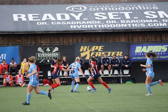 Players engage in a competitive soccer game on a field while others are seated on the bench watching attentively. The action is taking place in front of a wooden structure displaying multiple advertisements, including orthodontics, lacrosse, and field hockey.