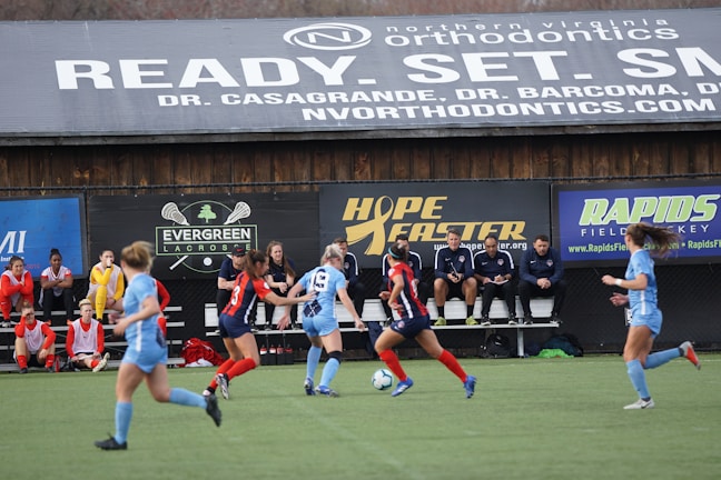 Players engage in a competitive soccer game on a field while others are seated on the bench watching attentively. The action is taking place in front of a wooden structure displaying multiple advertisements, including orthodontics, lacrosse, and field hockey.