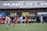 Players engage in a competitive soccer game on a field while others are seated on the bench watching attentively. The action is taking place in front of a wooden structure displaying multiple advertisements, including orthodontics, lacrosse, and field hockey.