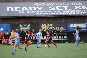 Players engage in a competitive soccer game on a field while others are seated on the bench watching attentively. The action is taking place in front of a wooden structure displaying multiple advertisements, including orthodontics, lacrosse, and field hockey.