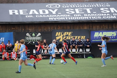Players engage in a competitive soccer game on a field while others are seated on the bench watching attentively. The action is taking place in front of a wooden structure displaying multiple advertisements, including orthodontics, lacrosse, and field hockey.