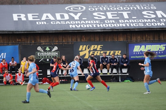Players engage in a competitive soccer game on a field while others are seated on the bench watching attentively. The action is taking place in front of a wooden structure displaying multiple advertisements, including orthodontics, lacrosse, and field hockey.