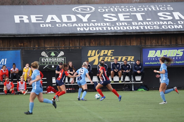 Players engage in a competitive soccer game on a field while others are seated on the bench watching attentively. The action is taking place in front of a wooden structure displaying multiple advertisements, including orthodontics, lacrosse, and field hockey.