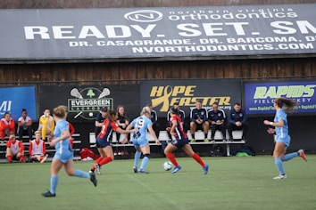 Players engage in a competitive soccer game on a field while others are seated on the bench watching attentively. The action is taking place in front of a wooden structure displaying multiple advertisements, including orthodontics, lacrosse, and field hockey.