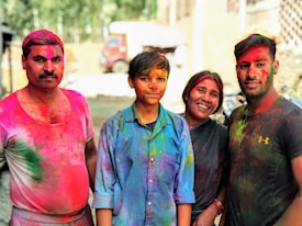 Four people are smiling and covered in vibrant colors, suggesting they are celebrating a festival involving colored powders. The background features an outdoor setting with some blurred vehicles and buildings.