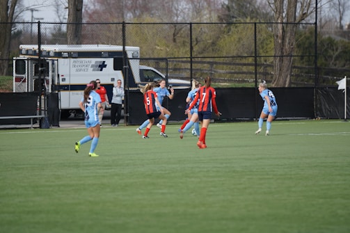 A group of female soccer players is actively engaged in a match on a green field. Some players are wearing blue uniforms, while others are in red and black striped jerseys. An ambulance is parked in the background near a fence, indicating a secure and prepared environment. Trees and additional fencing are visible around the field.