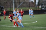 Players are actively engaging in a competitive soccer match on a green field with a black fence in the background. Two players in contrasting uniforms are prominently featured, vying for control of the soccer ball while other players are scattered in the background.