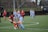 Players are actively engaging in a competitive soccer match on a green field with a black fence in the background. Two players in contrasting uniforms are prominently featured, vying for control of the soccer ball while other players are scattered in the background.