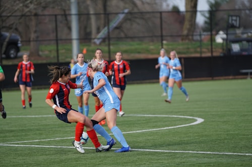 Players are actively engaging in a competitive soccer match on a green field with a black fence in the background. Two players in contrasting uniforms are prominently featured, vying for control of the soccer ball while other players are scattered in the background.