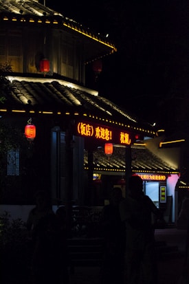 A traditional Asian building is illuminated at night with brightly glowing red lanterns and neon signage in Chinese characters. The roof features curved tiles with warm lighting outlining its structure. Shadows obscure some details, adding a mysterious touch.