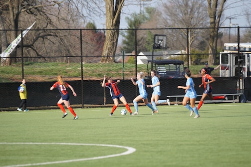 A group of female soccer players is actively engaged in a game on a green field, with two teams in different uniforms: light blue and dark blue with red accents. Nearby, a child wearing a yellow vest stands holding a soccer ball. The background features a black fence, leafless trees, and some park or neighborhood scenery.